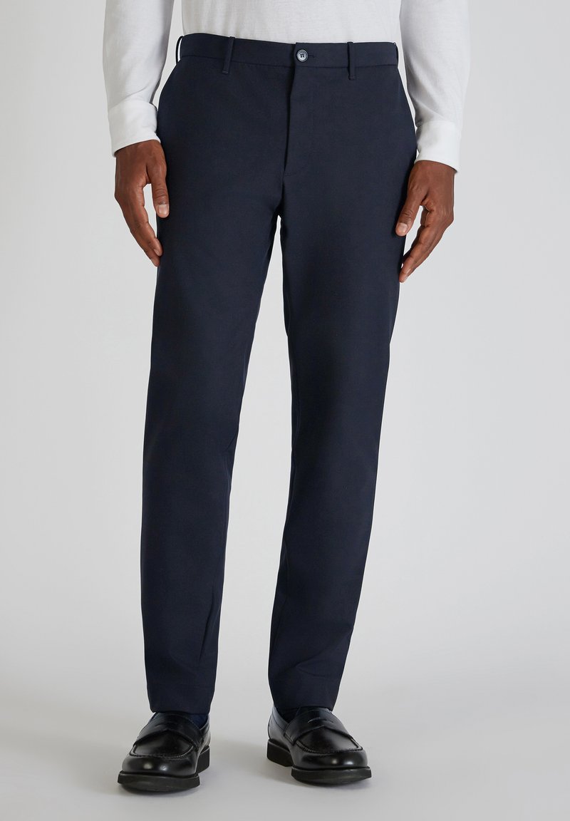 Marineblaue Baumwollhosen mit geradem Bein, Bund mit Knopfverschluss und Seitentaschen. Kombiniert mit schwarzen Slippern und einem weißen Langarmshirt.