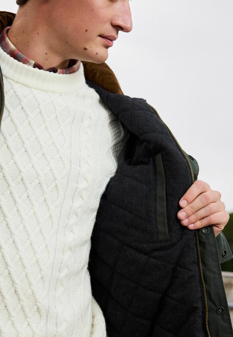 Gestrickter, cremefarbener Pullover mit Kabelmuster, unter einer dunkelgrünen Jacke mit braunem Futter. Das Innere der Jacke ist gesteppte und grau mit einer glatten Textur.