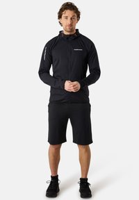 Schwarze Sportjacke mit Reißverschluss und Kapuze; verfügt über orangefarbene Akzente und reflektierende Details. Kombiniert mit schwarzen Shorts und schwarzen Turnschuhen.