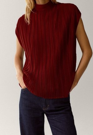 Woman wearing a sleeveless, high-neck, pleated dark red top tucked into dark blue jeans with hands in pockets.