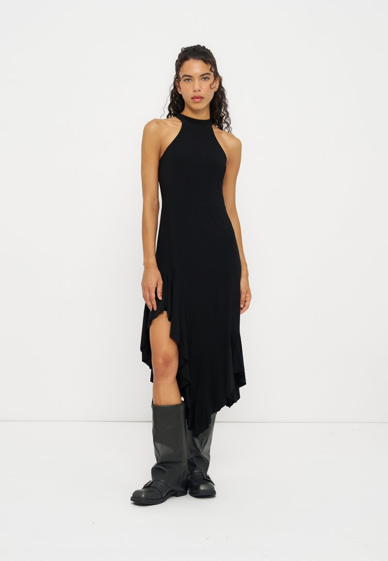 Femme aux cheveux bouclés portant une robe noire asymétrique sans manches et des bottes noires montantes sur un fond blanc uni.