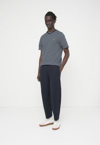 Μπλε ναυτικό ριγέ T-shirt με κίτρινο λογότυπο, σετ με φαρδιά μπλε παντελόνια και λευκά αθλητικά παπούτσια με καφέ σόλες. Απλός, καθημερινός σχεδιασμός.