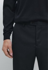 Mand iført sort langærmet skjorte og teksturerede sorte bukser, med venstre hånd hvilende ved siden af hoften mod en ensartet lys baggrund.