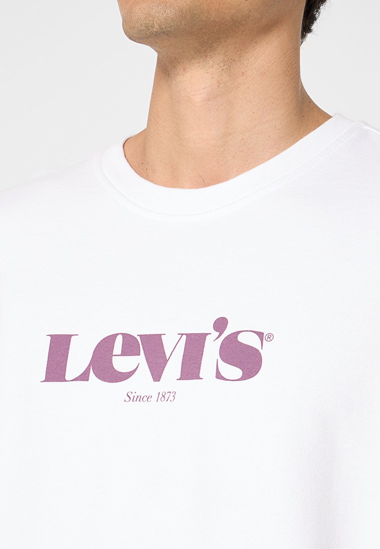 Camiseta blanca de algodón con cuello redondo, con un logo morado de Levi's y el texto "Desde 1873" debajo. Textura suave.
