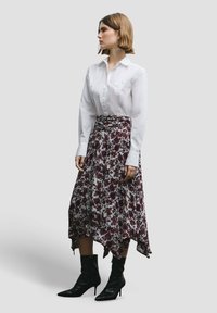 Witte blouse gecombineerd met een bloemen, asymmetrische rok met bordeauxrode en paarse patronen, aangevuld met zwarte hoge hakken.