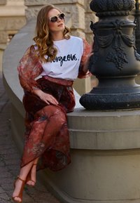 Czerwona, kwiatowa sukienka z przezroczystego materiału z długimi, zwiewnymi rękawami na białej bluzce z czarnym napisem, zestawiona z brązowymi sandałami na obcasie.