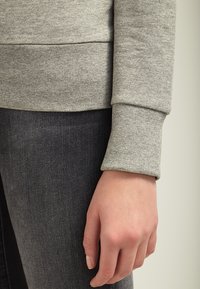 Felpa grigia chiara con polsini a costine, abbinata a jeans grigi scuri. Primo piano del braccio e della mano appoggiati sulla coscia.