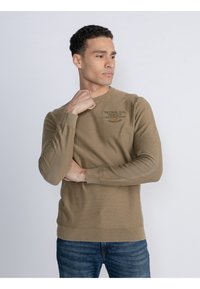 Khakifarbener Langarm-Sweatshirt mit Rundhalsausschnitt und gerippten Bündchen und Saum, mit gedrucktem Logo-Detail auf der Brust. Kombiniert mit blauen Jeans.