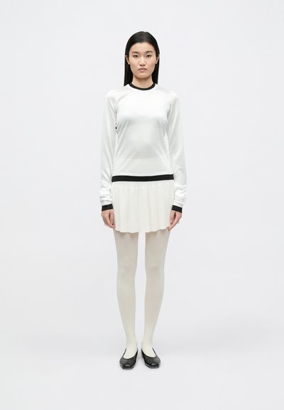 Haut blanc à manches longues avec bordure noire, associé à une jupe plissée couleur crème. Le mannequin porte des collants crème et des chaussures noires à enfiler. Design simple et moderne.