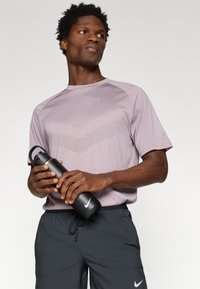 Ανοιχτό μοβ αθλητικό μπλουζάκι με γεωμετρικό σχέδιο και κοντά μανίκια, συνδυασμένο με μαύρες βερμούδες. Μοντέλο κρατάει μια μαύρη μπουκάλι νερού.