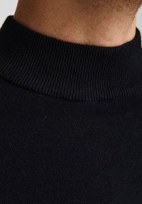Détail d'un pull en tricot noir avec un col montant côtelé, une texture lisse, et un gros plan sur le tissage fin du tissu.