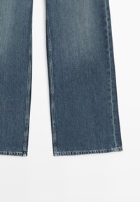 Jeans a gamba dritta in denim blu con cuciture visibili su sfondo bianco, che mostrano la metà inferiore delle gambe.