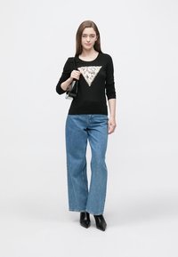 Schwarzes Langarmshirt mit einem Pailletten-Dreiecksmuster, kombiniert mit hochtaillierter blauer Denim-Jeans und schwarzen, hochhackigen Stiefeletten.