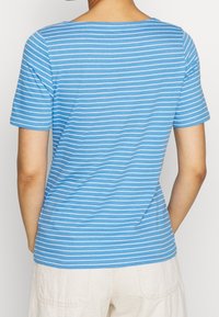 Persona vestida con una camiseta de manga corta a rayas horizontales en azul y blanco y pantalones de un color claro, vista desde atrás.