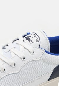 Bela teniska z zgornjim delom iz teksturirane usnje, modrimi akcenti na jezik in peti ter logotipom. Ima bele vezalke in okrogle luknje.