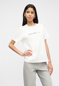 Νεαρή γυναίκα με μακριά σκούρα μαλλιά στέκεται με το ένα χέρι στη μέση, φοράει λευκό μπλουζάκι με το κείμενο «Σ' αγαπώ.» και ανοιχτόχρωμο γκρι τζιν.