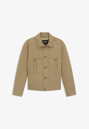 Vaaleanruskea puuvillatakki, jossa on kauluksellinen pääntie, pitkät hihat ja kaksi etutaskua, joissa on napilliset suljennat. Brändin merkki näkyy sisäpuolella.