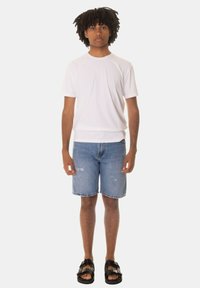 T-shirt bianco a maniche corte, pantaloni corti di jeans blu con orli sfrangiati e sandali neri. Il modello sta in posizione eretta su uno sfondo neutro.