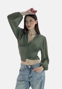 Olivgrüne Bluse mit tiefem V-Ausschnitt, langen gepufften Ärmeln und dekorativen Knöpfen vorne, kombiniert mit hellblauen, hoch taillierten Jeans.