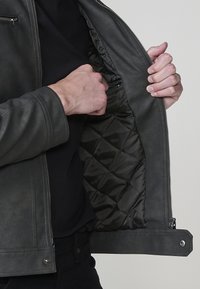 Giacca in pelle grigia con un rivestimento interno trapuntato scuro. Presenta una cerniera frontale e una tasca con cerniera sul lato sinistro. Vestibilità su misura e texture liscia.