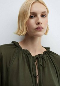 Bluză de culoare verde măslinie cu decolteu rotund, detalii pliate și închidere printr-o fundă. Material moale, ușor, cu o textură ușor transparentă.