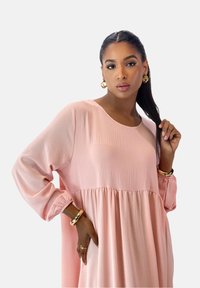 Robe rose pâle à manches longues avec un col arrondi, présentant une taille froncée et un tissu texturé. Des bracelets dorés ornent le poignet.