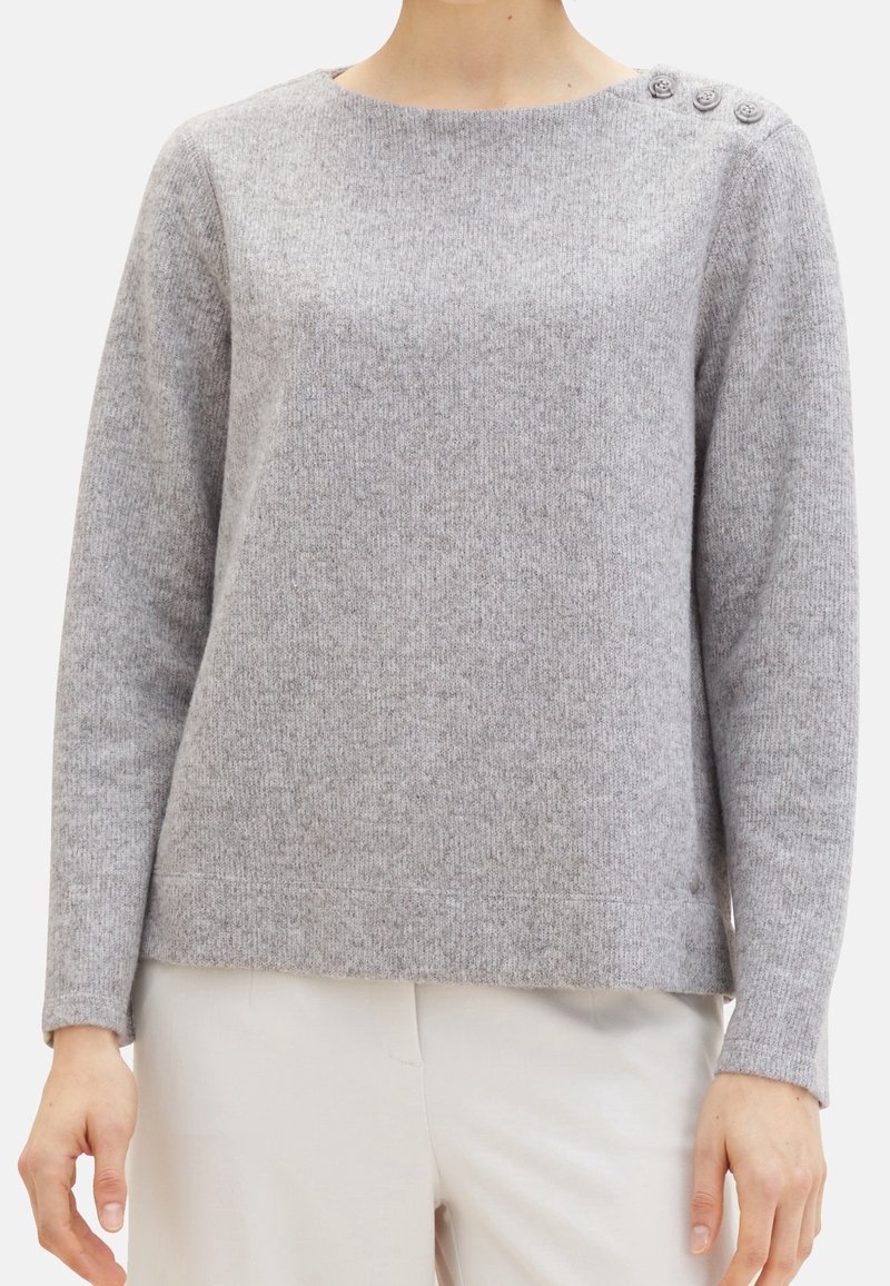 Femme portant un pull en maille gris clair avec trois boutons sur l'épaule gauche, associé à un pantalon beige clair.