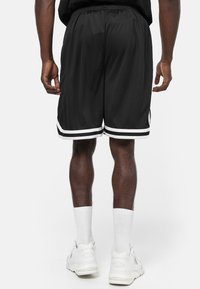 Bermudas deportivas negras con franjas laterales blancas, cinturilla elástica y tejido ligero. Combinadas con calcetines blancos y zapatillas.