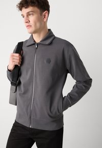 Giacca grigia con zip e colletto, tessuto texturizzato e logo esagonale sul petto. Abbinata a pantaloni scuri e una borsa a tracolla.