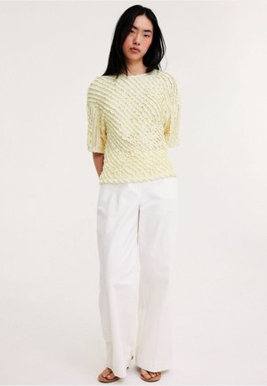 Frau trägt ein strukturiertes gelbes kurzärmeliges Oberteil, weite weiße Hosen und braune offene Sandalen, steht vor einem schlichten Hintergrund.