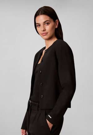 Femme aux cheveux foncés portant une veste noire boutonnée sur un haut noir et un pantalon noir, la main dans la poche, sur un fond gris uni.