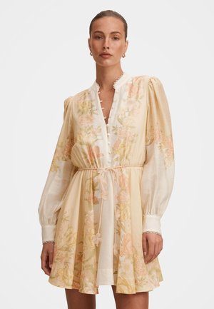 Γυναίκα που φοράει μίνι φόρεμα κρεμ με φλοράλ σχέδιο, μακριά φουσκωτά μανίκια, κουμπιά στο εμπρός μέρος, γιακά με κυματιστό τελείωμα και ζώνη στη μέση δεμένη με φιόγκο.