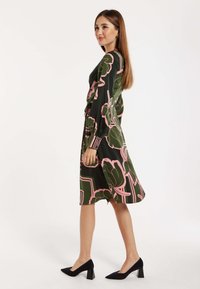 Floral dress in dark green with pink and black patterns, long sleeves, waist tie, and a midi length, paired with black block-heeled shoes.