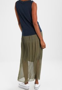 Kvinna i ärmlös marinblå topp, olivgrön genomskinlig plisserad maxikjol och vita platåskor, står framför en enkel vit bakgrund.