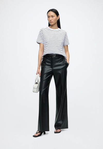 Femme portant un T-shirt rayé blanc et noir, un pantalon en cuir noir à jambes larges, des sandales à talons noires, et tenant un petit sac à main blanc.