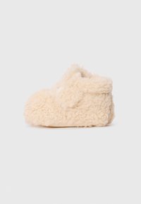 Beige, fluffy tøfler laget av myk lammeskinnsmateriale med avrundet form og subtile sømdetaljer på siden.