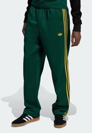 Mężczyzna ubrany w zielone dresy Adidas w cienkie paski z żółtymi paskami po bokach oraz czarne buty sportowe Adidas z białymi paskami.