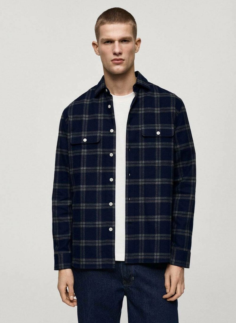 Camicia di flanella blu marina con motivo a quadri grigi, chiusura con bottoni sul davanti, due tasche sul petto e colletto a punta. Indossata sopra una maglietta bianca.