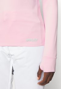 Close-up of person wearing a light pink long-sleeve top with thumbhole and white pants, showing logo near hem on the left side.