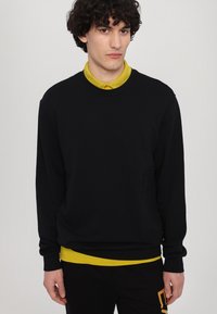 Felpa nera con collo a giro e maniche lunghe, sovrapposta a una camicia gialla brillante con colletto. Presenta un dettaglio con tasca frontale.