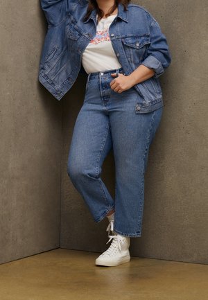 Person trägt blaue Jeansjacke und Jeans mit weißen Sneakers, steht an einer grauen Wand- und Bodenecke.
