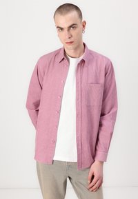 Light pink button-up shirt with a pocket and vertical stripes, worn over a white t-shirt. The fabric appears textured, with long sleeves.