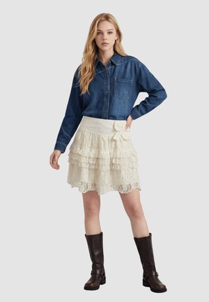 Mujer joven con camisa vaquera azul, falda de encaje cremosa en capas con lazo y botas altas hasta la rodilla de color marrón oscuro, de pie frente a un fondo liso.