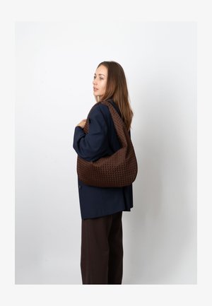 Hnědá tkaná kabelka s texturovaným povrchem a měkkým tvarem. Nositelná přes rameno, kombinovaná s tmavě modrým kabátem a tmavými kalhotami.