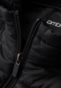 Chaqueta acolchada negra con cuello alto, que presenta una cremallera visible en un acabado mate bronce y una etiqueta de la marca que dice "ombre".