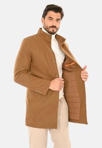 Cappotto in lana marrone con collo alto e chiusura con bottoni, caratterizzato da una tasca interna a contrasto e fodera trapuntata. Vestibilità sartoriale e design pulito.