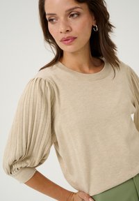 Beige knit sweater with short, pleated sleeves and a round neckline. The fabric has a smooth texture, paired with green trousers.