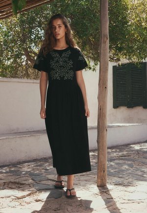 Kobieta z długimi kręconymi włosami w czarnej sukience maxi z białą kwiatową haftowaną dekoracją, stojąca na zewnątrz na kamiennej ścieżce obok drewnianego słupa.