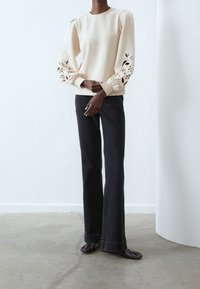 Bluza w kolorze kremowym z bufkami na rękawach i wycięciami w kształcie kwiatów, zestawiona z rozszerzanymi ciemnymi dżinsami i brązowymi płaskimi butami.