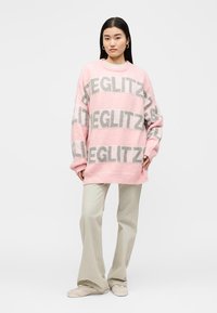 Νεαρή γυναίκα με ροζ πουλόβερ σε μεγάλο μέγεθος με γκρι κείμενο "GLEGLITZ", μπεζ παντελόνια με φαρδιά πόδια και ανοιχτό μπεζ αθλητικά παπούτσια σε λευκό φόντο.
